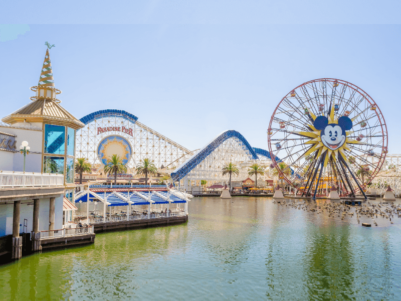 A vibrant amusement park scene featuring a large Ferris wheel with a Mickey Mouse face, a blue and white roller coaster, palm trees, and a lake in the foreground under a clear blue sky.