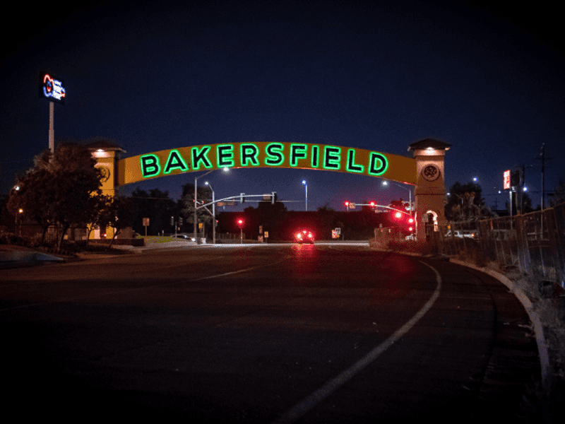 A large yellow arch with green illuminated letters spelling “Bakersfield” spans a roadway at night, with streetlights, traffic signals, and a few cars visible in the background.