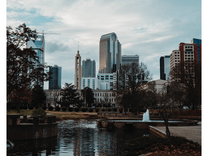 City skyline with modern tall buildings in the background, a park with trees and a pond in the foreground, and a small fountain near the water, under a cloudy sky.