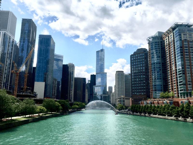 A sunny view of the Chicago River with turquoise water, modern skyscrapers on both sides, trees lining the riverbank, and a water fountain spraying in the center under a partly cloudy sky.