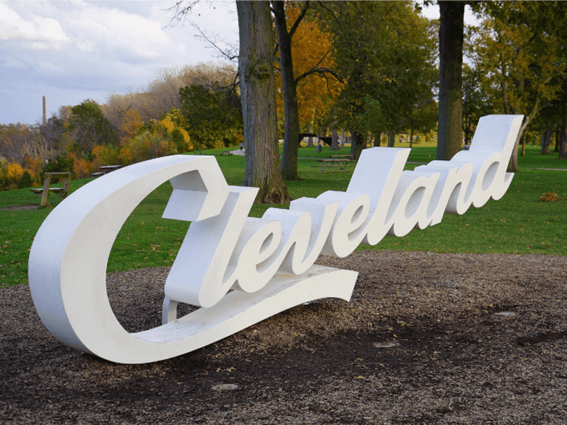 A large white script sign that reads Cleveland sits in a park with green grass, trees, and autumn foliage in the background.