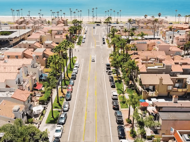 Aerial view of a coastal neighborhood with palm trees, houses lining both sides of a straight road, parked cars, and a sandy beach with the ocean in the background.