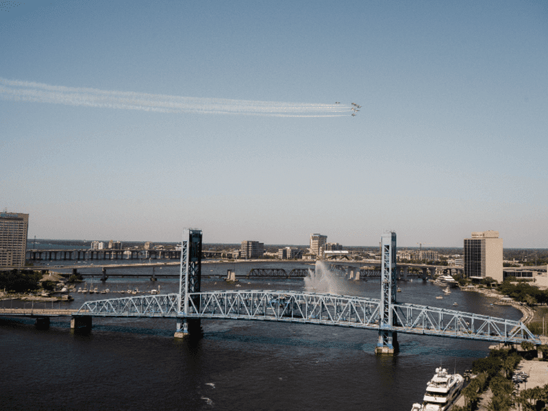A blue drawbridge spans a wide river in a city, with jets flying in formation and leaving white trails in the clear sky above. Buildings and boats line the riverbanks.