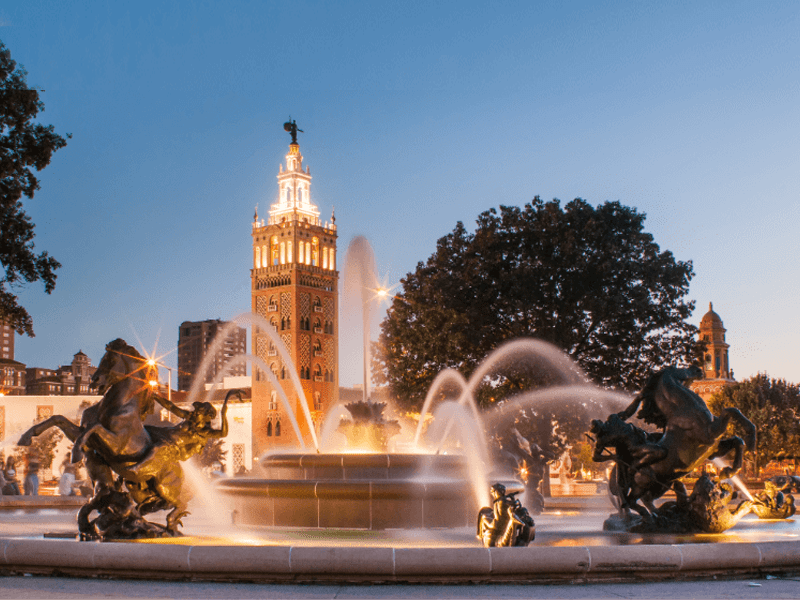 A large fountain with bronze horse statues sprays water, set against a dusk sky. In the background, a tall, illuminated tower and surrounding buildings are visible, with trees framing the scene.