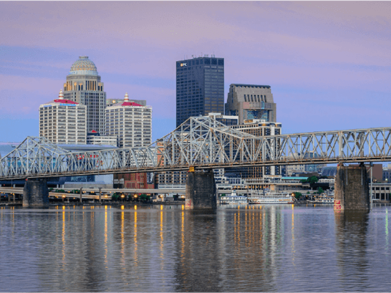 A cityscape view of downtown Louisville, Kentucky, featuring a steel truss bridge over the Ohio River with tall buildings and a pink and purple sky in the background.
