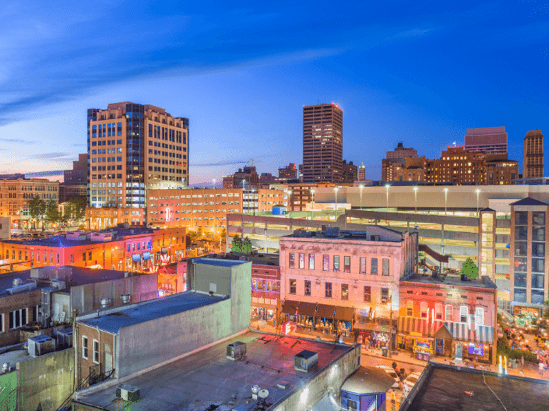 Downtown Shreveport, Louisiana at dusk, showing historic buildings with colorful lights, a multi-level parking garage, and modern office towers under a blue evening sky.