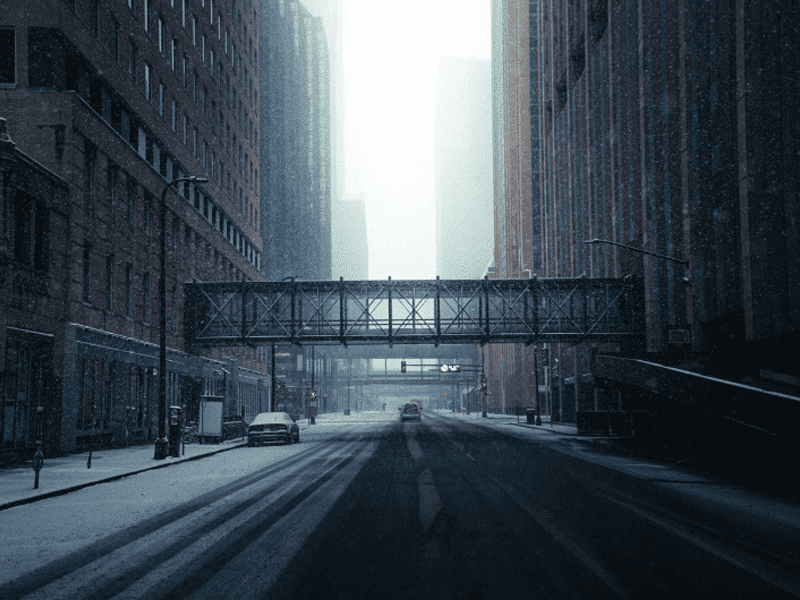 A snowy, empty city street is lined with tall buildings and two enclosed pedestrian bridges span across the road. A few parked cars and faint tire tracks are visible on the snow-covered pavement.