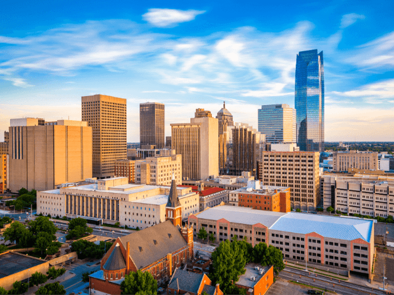 Downtown cityscape with modern skyscrapers, historic buildings, and a church in the foreground, under a blue sky with wispy clouds.