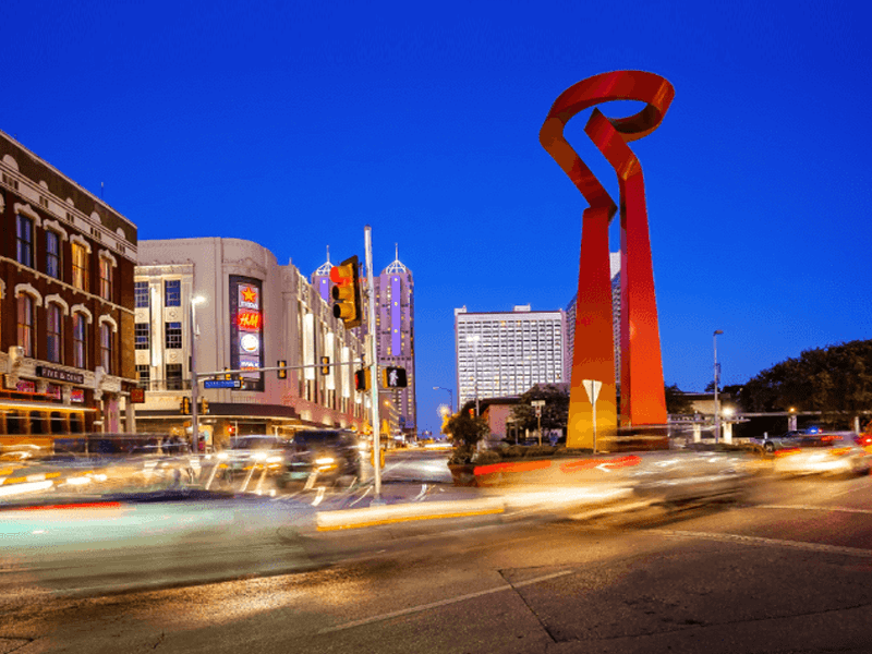 A city street at dusk features the Torch of Friendship sculpture, surrounded by blurred car lights, historic buildings, and modern hotels under a deep blue sky in downtown San Antonio, Texas.