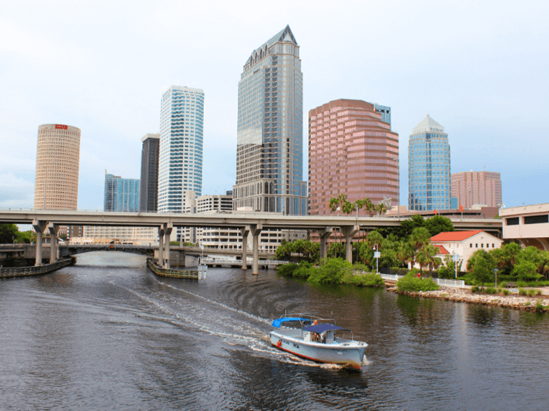 A small boat moves along a river in front of a city skyline with tall modern buildings and a raised bridge on a cloudy day.