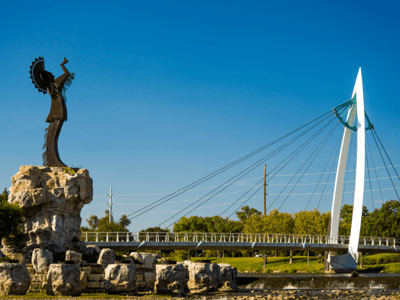 A tall statue of a Native American figure stands on a rocky pedestal next to a modern cable-stayed pedestrian bridge, with trees and a clear blue sky in the background.