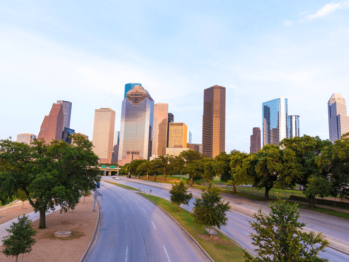 A wide, empty road leads toward the modern downtown skyline of Houston, Texas, with tall glass buildings surrounded by green trees under a clear blue sky.