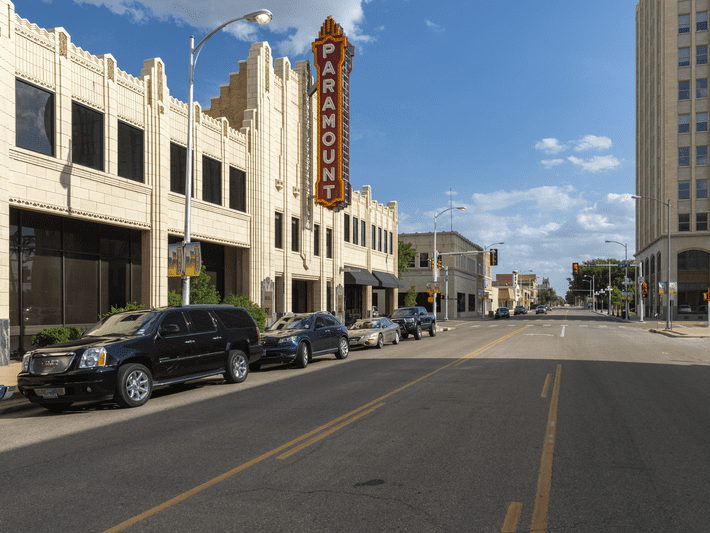 A street view of a city with parked cars along the curb. A vintage Paramount theater sign is attached to an art deco-style building on the left, with blue sky and few clouds above.