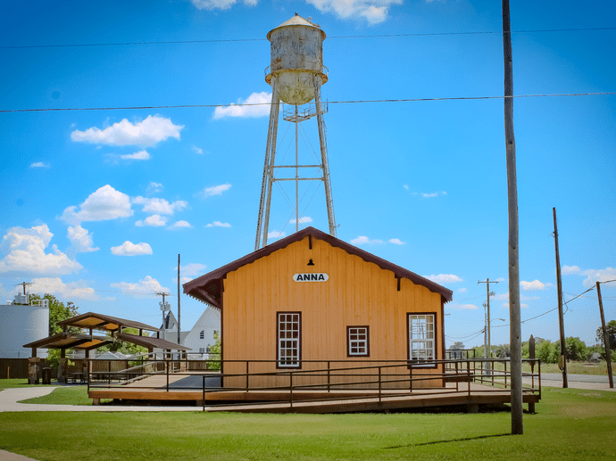 A small, yellow wooden building with a sign reading Anna stands in front of a tall, rusty water tower under a bright blue sky with scattered clouds. Power lines and grassy areas surround the scene.