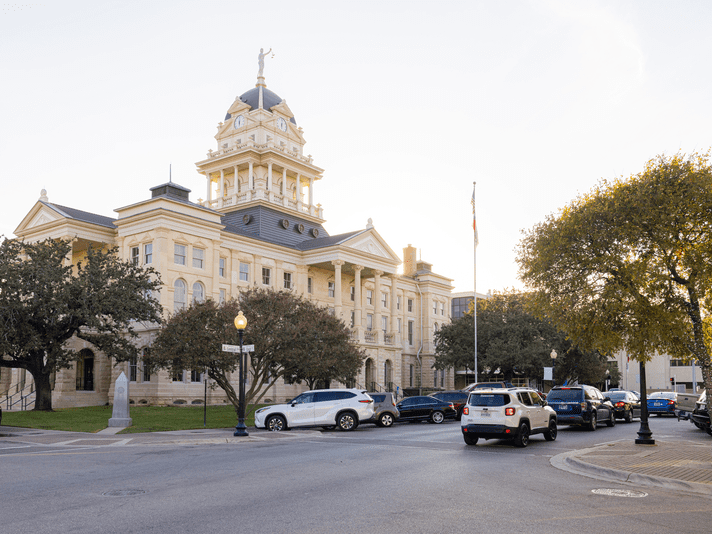 Historic courthouse with a dome and statue on top, surrounded by trees and parked cars along the street. The sky is clear and a flagpole is visible near the entrance.