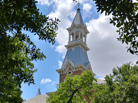 A tall, pointed clock tower with a blue-gray roof and white trim rises above green trees, framed by leafy branches against a partly cloudy blue sky.
