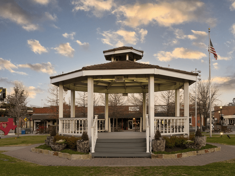 A white wooden gazebo stands in the center of a park with an American flag nearby, surrounded by shops and trees under a cloudy sky at sunset.