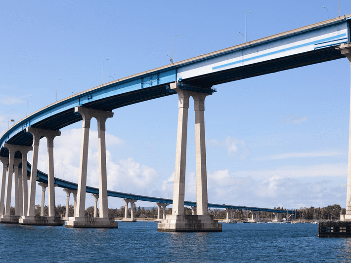 A tall, curved bridge with blue accents supported by concrete pillars stretches over a body of water under a clear blue sky. Trees and buildings are visible in the background.