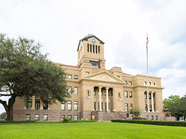 A historic courthouse with a clock tower, American flag, large columns, and wide steps, surrounded by neatly trimmed grass and trees under a cloudy sky.