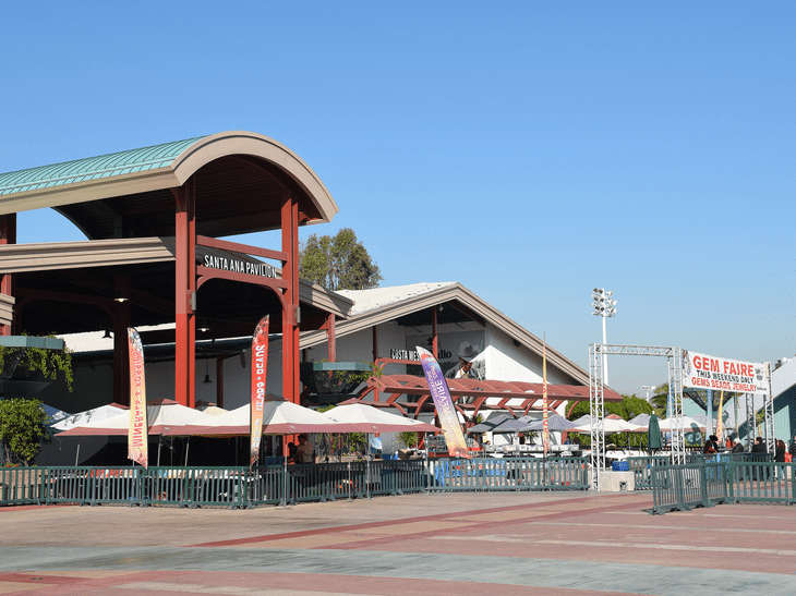 Outdoor scene of Santa Ana Pavilion with red and white structures, event tents, colorful banners, and a large GEM FAIRE sign. The area is fenced and mostly empty, with clear blue sky overhead.