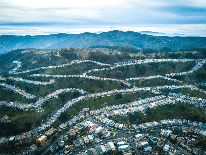 Aerial view of a hilly suburban neighborhood with winding roads and rows of houses, set against green hills and cloudy skies in the background.