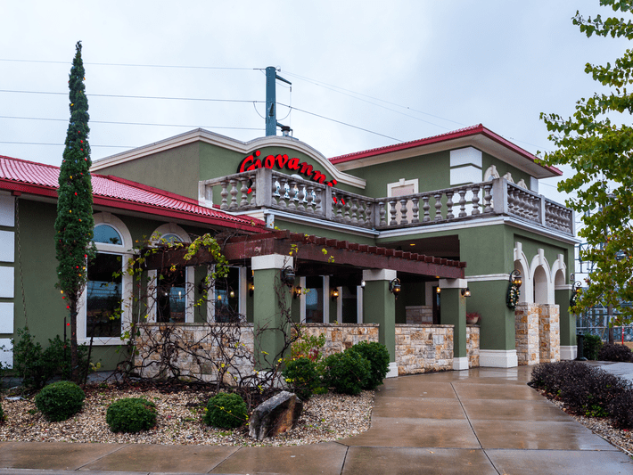 A green and beige restaurant building with a red roof, stone accents, arched windows, and a balcony. The sign Giovannis is on top, and the sidewalk and landscaping appear wet from recent rain.