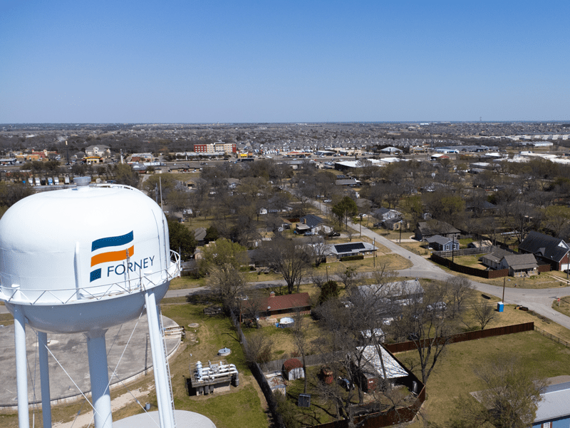 A white water tower labeled FORNEY stands in the foreground, overlooking a suburban neighborhood with houses, trees, and a sprawling urban landscape under a clear blue sky.