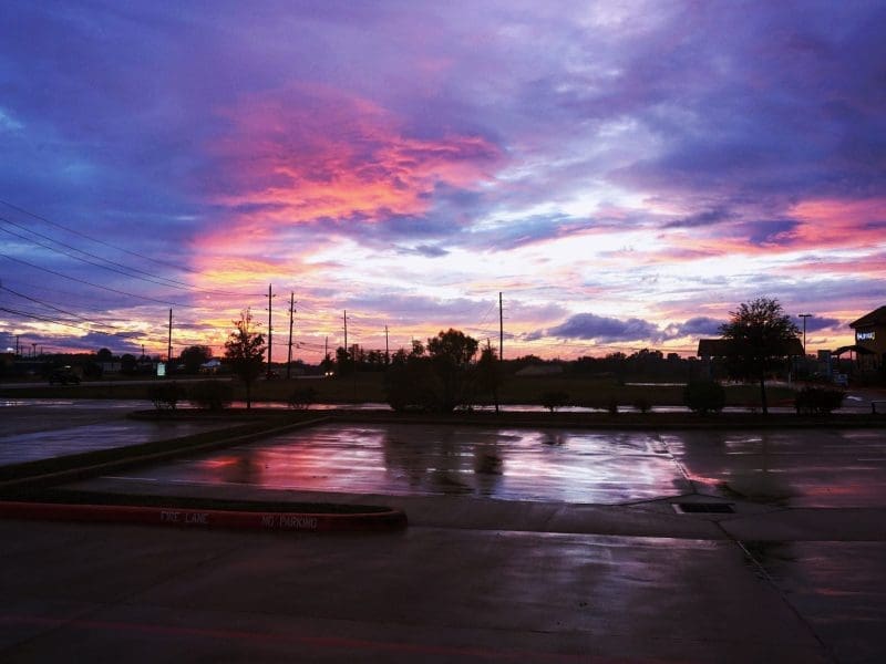 A mostly empty parking lot with wet pavement reflecting a dramatic sunrise or sunset sky filled with vibrant pink, purple, and blue clouds. Power lines and a few trees are visible in the background.