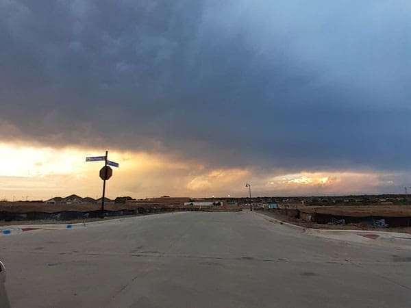 A wide, empty street in a suburban area under a dramatic sky, with dark clouds on one side and a glowing sunset on the horizon. A street sign and stop sign stand at the intersection.
