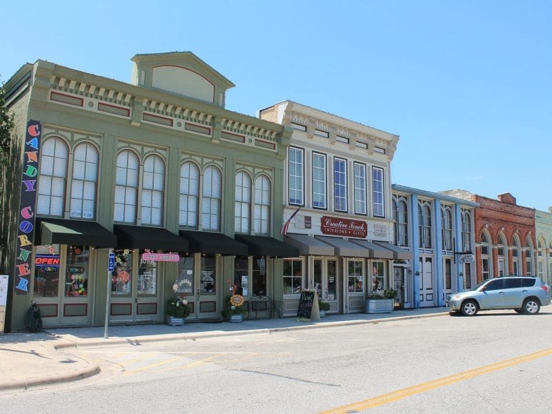 A row of colorful, historic storefronts lines a sunny small-town street. One shop displays a vertical Candy Corner sign, and a silver car is parked along the curb. The sky is clear and blue.