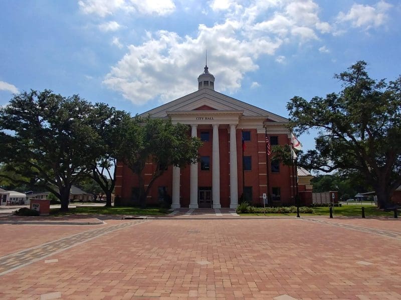 Red brick city hall with white columns and a small cupola on top, surrounded by trees and a large brick courtyard under a partly cloudy sky.