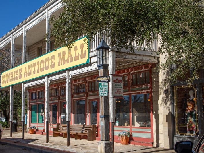 A historic two-story building with a large yellow “Sunrise Antique Mall” sign, red-trimmed windows, benches out front, and a streetlamp with parking signs in a sunny, tree-lined area.