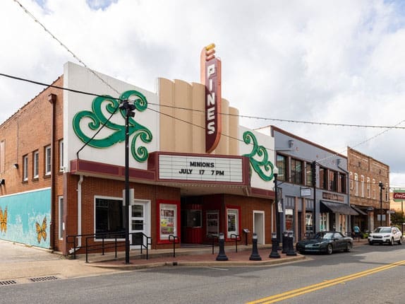 A vintage theater with a marquee reading “MINIONS JULY 17 7PM” sits on a street corner. The building has art deco details, a vertical sign that says Pines, and neighboring storefronts. A mural is visible on the left wall.