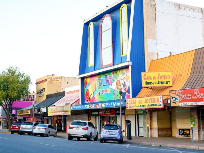 A colorful row of shops with bright awnings and signs, including jewelry stores and a party supply store, lines a street with parked cars and a few trees.