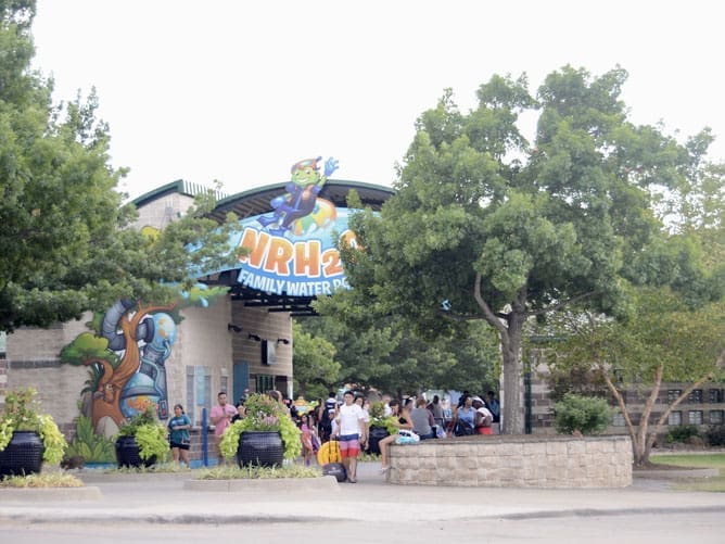 Entrance to NRH2O Family Water Park with people walking in, trees and plants around, and a colorful sign featuring a frog-shaped mascot above the entrance.