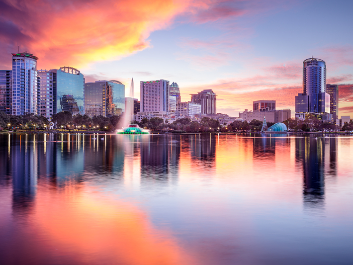 Orlando city skyline at sunset with colorful clouds, modern buildings, and a fountain reflecting on calm lake water in the foreground.