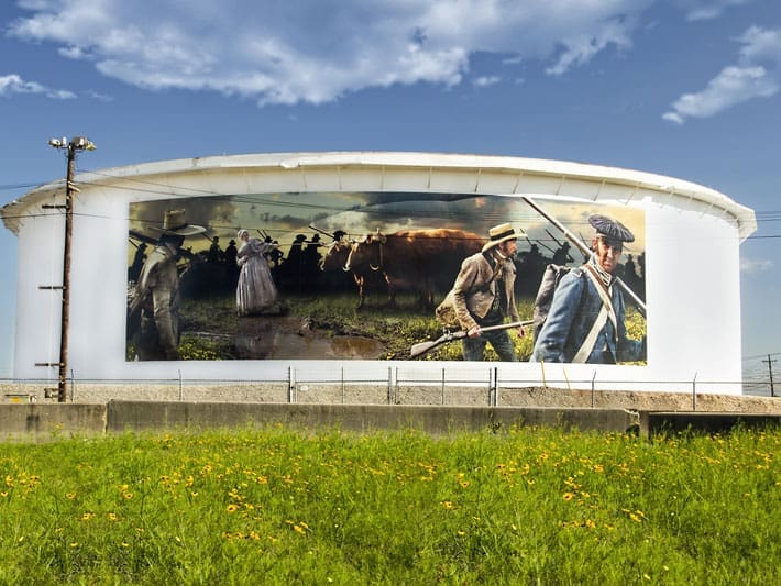 A large cylindrical white building displays a mural of pioneers and cattle crossing a muddy field, with a woman in a bonnet and men in hats leading the way. Yellow wildflowers grow in the grassy area in front.