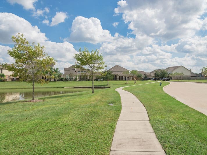 A curved sidewalk runs through a grassy suburban area with young trees, a small pond on the left, and houses in the background under a partly cloudy sky.