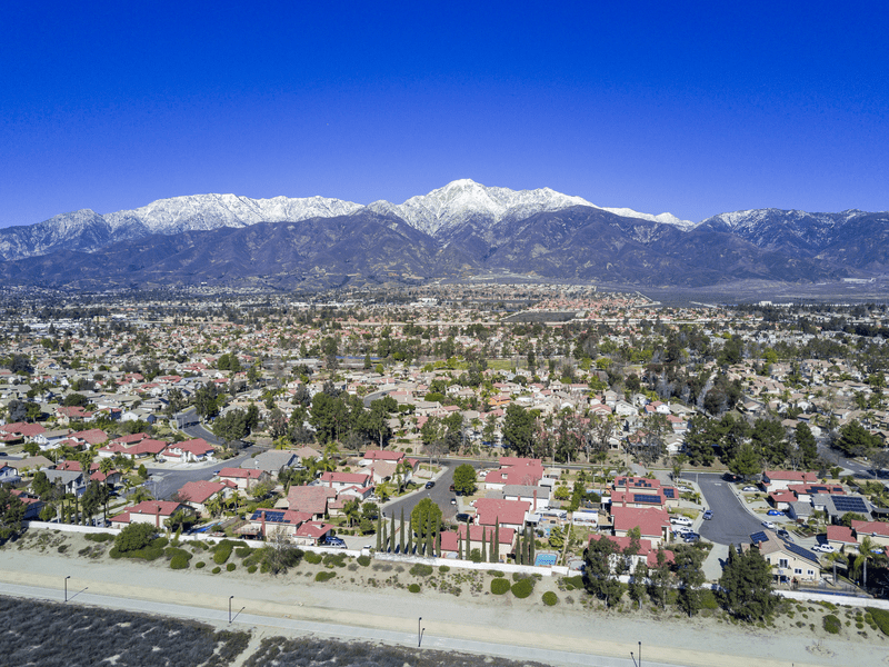 Aerial view of a suburban neighborhood with red-roofed houses, green trees, and a clear road in the foreground, set against a backdrop of mountains with snow-capped peaks under a bright blue sky.