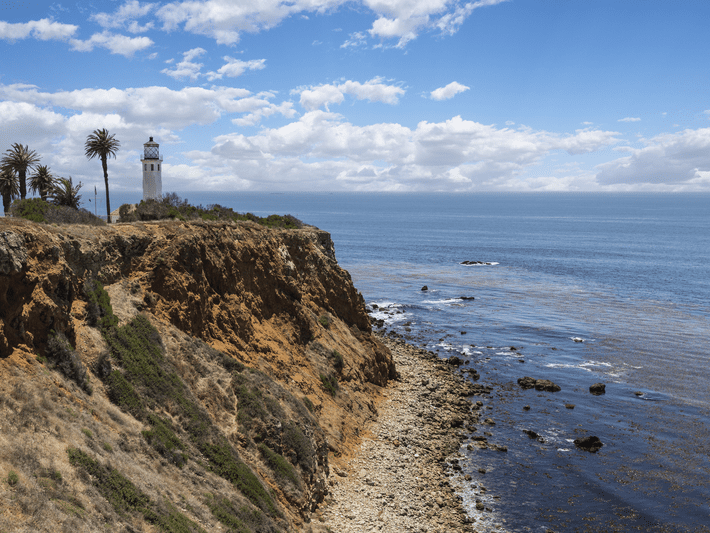 A white lighthouse stands on a rocky, brown cliff overlooking the ocean, with palm trees nearby and scattered clouds in a blue sky. The coastline below is covered with rocks and bordered by calm water.