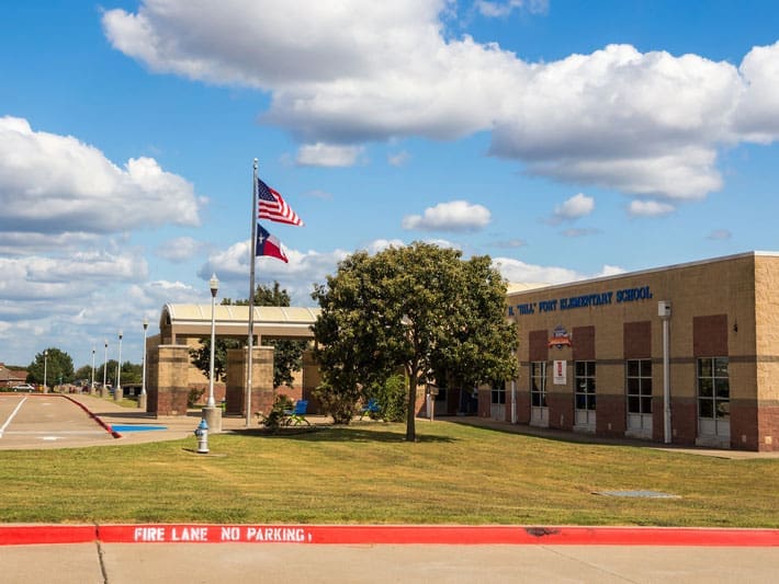 A single-story brick elementary school building with an arched entrance, flagpoles displaying the US and Texas flags, and a grassy lawn under a partly cloudy sky. FIRE LANE NO PARKING is painted on the curb.
