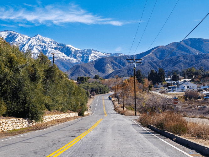 A rural road with yellow lines curves through a landscape of green shrubs and utility poles, leading toward snow-capped mountains under a clear blue sky. Modest buildings and a stop sign appear on the right side.