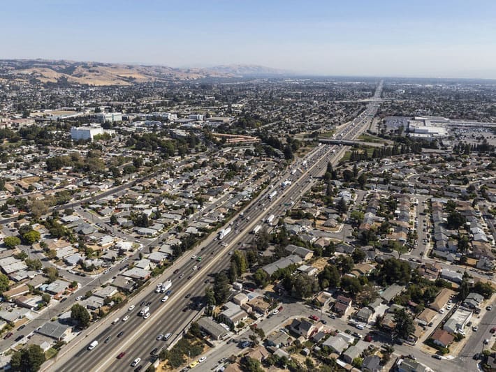Aerial view of a sprawling suburban neighborhood with a busy freeway running through the center, surrounded by houses, buildings, and distant hills under a clear sky.