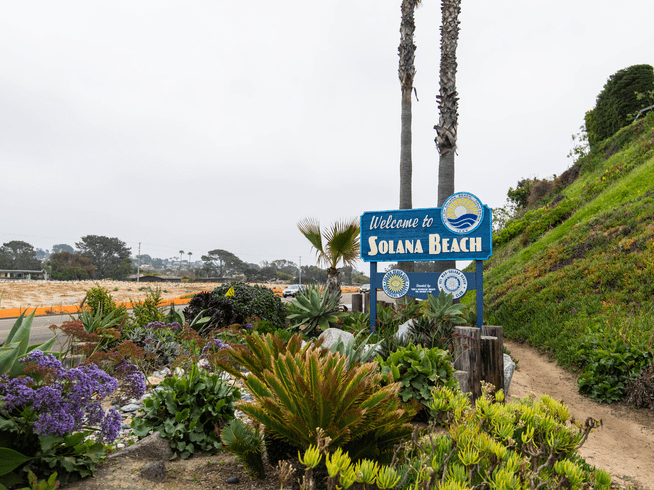 A blue Welcome to Solana Beach sign stands among lush plants and flowers beside a sandy path, with palm trees and a cloudy sky in the background.