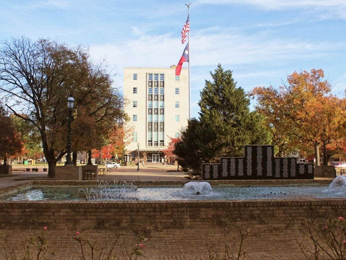 A brick fountain with water jets sits in front of a tall building with Texas and U.S. flags. Trees with autumn leaves surround the plaza under a blue sky.