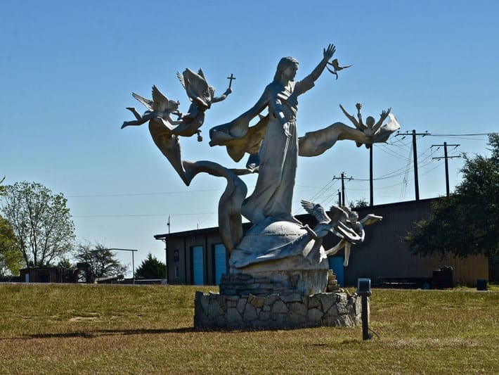 A large outdoor sculpture of a robed figure with outstretched arm surrounded by flying birds and angels, set on a stone base in a grassy area with buildings and power lines in the background.