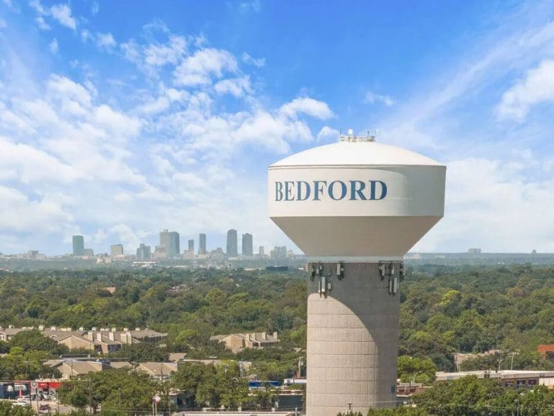 A large white water tower labeled BEDFORD stands above a suburban landscape with trees and houses, with a city skyline visible in the distance under a partly cloudy blue sky.