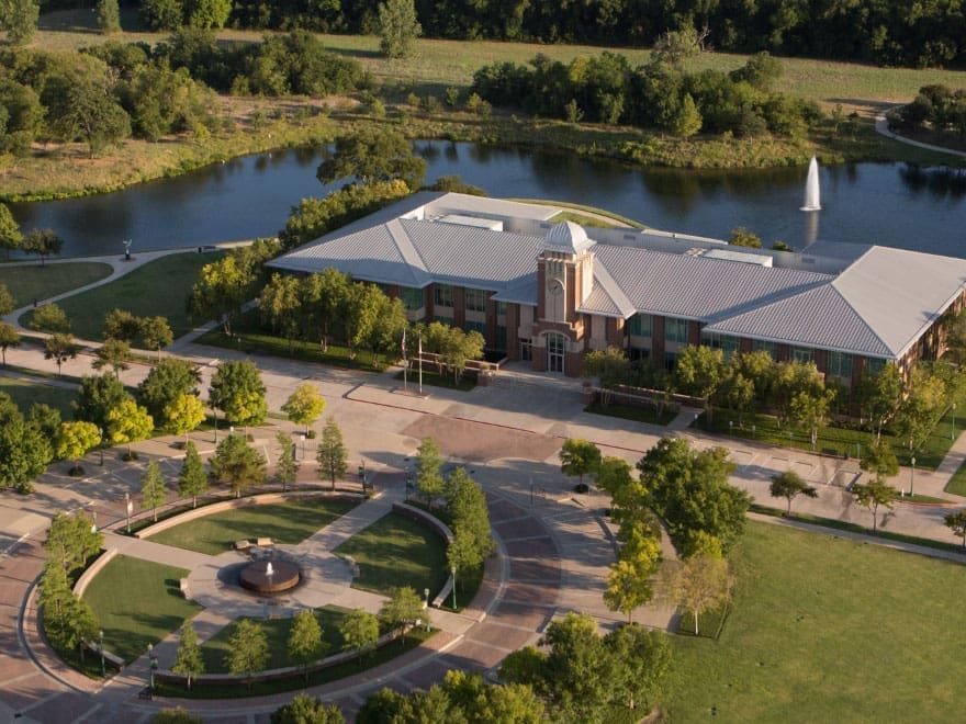 Aerial view of a large building with a clock tower, surrounded by trees, a circular plaza with a fountain, and a pond with a fountain in a park-like setting.