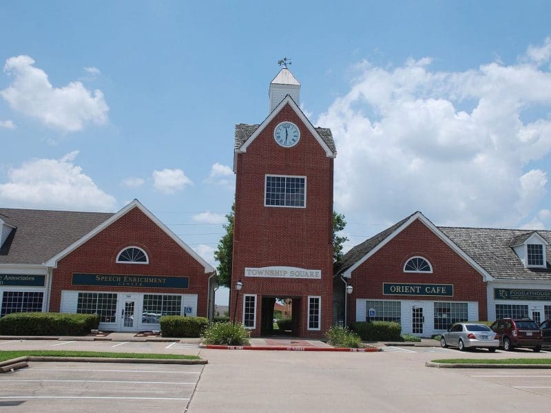 A red-brick building with a central clock tower labeled “Township Square” is flanked by a “Speech Enrichment” office and “Orient Cafe.” Several cars are parked in front under a blue, partly cloudy sky.